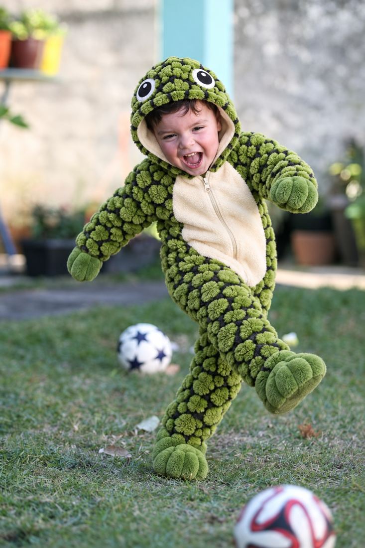 Menino rindo, de macacão de tartaruga, chutando uma bola no jardim